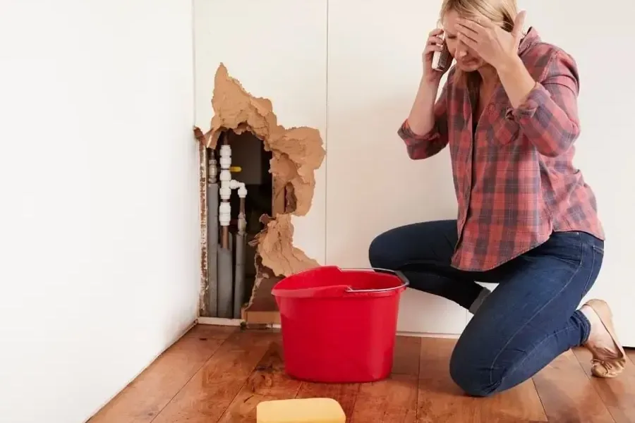 Wall opened around leaking supply pipes with a bucket on a wet wood floor